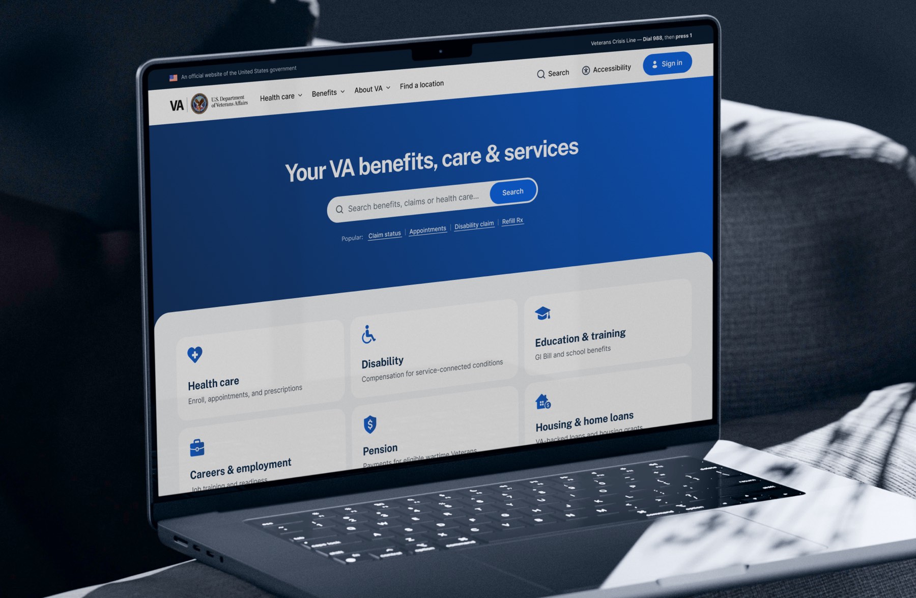 The redesigned VA.gov homepage open on a laptop: a deep blue hero reading “Your VA benefits, care & services,” one centred search field, a row of popular task links, and a grid of six benefit cards below — health care, disability, education and training, careers and employment, pension, and housing and home loans.